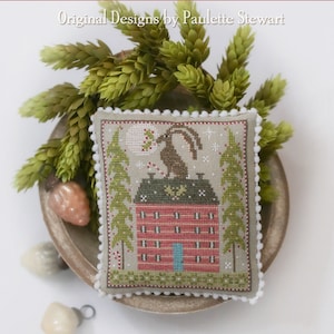 May include: A cross-stitch sampler featuring a red house with a blue door, a rabbit on the roof, and a full moon in the sky. The sampler is titled "Christmas Mini Moon" and is by Plum Street Samplers. Original Designs by Paulette Stewart.