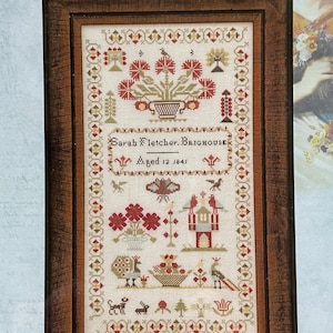 May include: A framed cross-stitch sampler with a white background and colorful embroidery. The sampler features a floral design, a house, and a bird. The text "Sarah Fletcher, Brighouse Aged 12 1841" is embroidered in the center of the sampler.