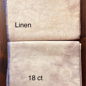 May include: Two folded linen fabric pieces. The top piece is a light beige color with the word "Linen" printed on it. The bottom piece is a light beige color with the text "18 ct" printed on it. Both pieces have a blue stitched edge.