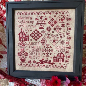 May include: A framed cross-stitch sampler with a cream-coloured background and red embroidery. The sampler features an alphabet, numbers, and various designs including a house, a rabbit, and a flower pot.