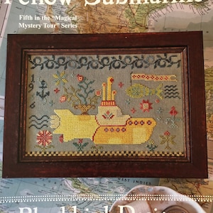 May include: A cross-stitch pattern featuring a yellow submarine with a red periscope and a fish swimming above it. The pattern is framed in a dark wood frame and is surrounded by a map with the text "Yellow Submarine" and "Blackbird Designs".