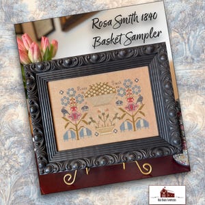 May include: A framed cross-stitch sampler, labelled "Rosa Smith 1840 Basket Sampler." The design includes a central basket motif with floral and bird elements in blue, red, and beige, encased in a dark, decorative frame.