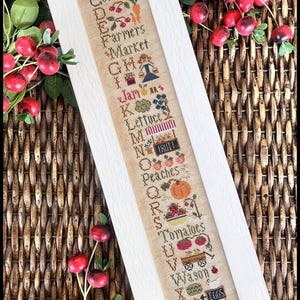 May include: A white framed cross-stitch sampler with an alphabet design featuring farm-themed words and images. The sampler includes letters A through Z with words like "Corn", "Farmers Market", "Jam", "Lettuce", "Peaches", "Tomatoes", "Wagon", and "Eggs".