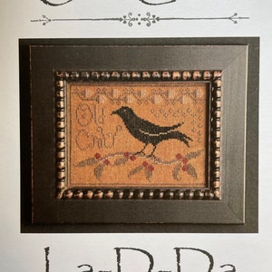 May include: A framed cross-stitch sampler with the text "Old Crow" at the top. The sampler features a black crow perched on a branch with red berries. The frame is dark brown, and the background is tan.