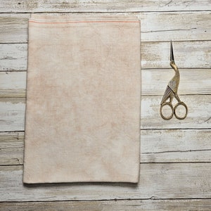 May include: A beige linen fabric with a subtle, uneven texture. The fabric is folded in half and laid flat on a whitewashed wooden surface. A gold-toned bird-shaped pair of scissors is placed to the right of the fabric.