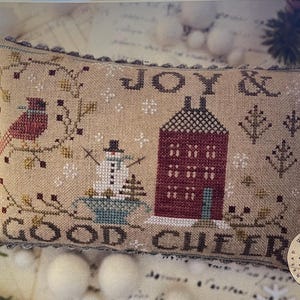 May include: A beige, rectangular, cross-stitched pillow with the words "JOY & GOOD CHEER". The design includes a red bird, a snowman, a house, and floral elements. The pillow has a decorative border.