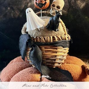 May include: A wicker basket pincushion with a black ribbon, decorated with a white ghost, a black cat, a pumpkin, and a skull. The pincushion is labeled "Witch's Tools" and "Pins and Fobs Collection 2024 Needlework Marketplace Collection by The Primitive Hare".