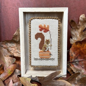 May include: A framed cross-stitch design featuring a squirrel sitting on a pumpkin with a leaf above its head. The design is in shades of brown, orange, and white.