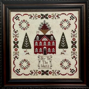 May include: Framed cross-stitch artwork featuring a red house with multiple windows, flanked by green trees. The design includes floral wreaths, decorative borders, and the text "Joy To The World." The piece is framed in a dark brown wooden frame.