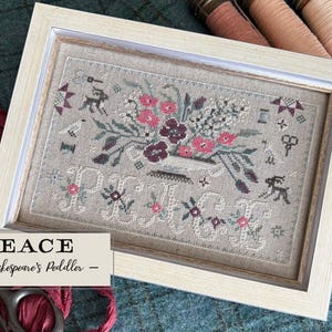 May include: Framed cross-stitch art featuring a floral arrangement in a vase, with the word "PEACE" stitched below. The artwork is in a light-colored frame. The background is a muted green fabric. Pink and burgundy flowers are the focal point.