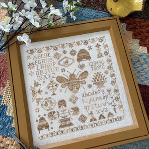 May include: A framed cross-stitch sampler with a bee and floral designs in brown and beige hues. The sampler includes the alphabet, numbers, and decorative elements. The wooden frame is a warm, golden brown colour.