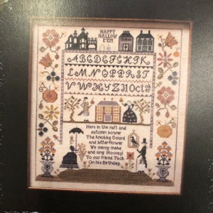May include: A cross-stitch pattern featuring a charming autumn scene with a house, a bird, and a couple. The pattern includes the text "Happy Hallow E'en" and the alphabet. The text below reads "Here in the ruft and autumn bower The knobby Gourd and bitter flower We merry-make and sing Hooray! To our friend Jack On his Birthday."
