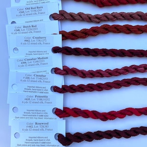 May include: A collection of 10 skeins of 12-strand silk embroidery floss in shades of red, from crimson to rosewood. Each skein is 6 yards long and hand-dyed in the USA. The labels include the color name, number, lot number, and length.