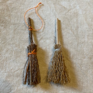 May include: Two miniature brooms made from natural materials. One broom has a brown wooden handle and is tied with orange string. The other broom has a light brown handle and is tied with white string.