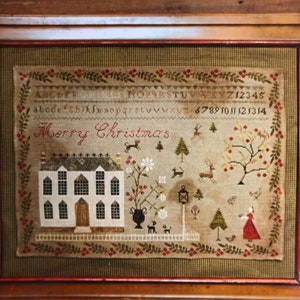 May include: A cross-stitch sampler with a white house, trees, and a woman in a red dress. The sampler is framed in a wooden frame with a green and brown plaid mat. The text "Merry Christmas" is stitched in black thread.