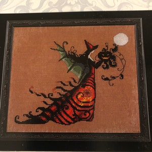 May include: Cross-stitch artwork in a black frame. The image features a witch with a pumpkin head, holding a moon. The witch wears a black hat and dress with orange and black details. Green bat wings are visible.