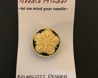 May include: A white needle minder with a black border and a floral design. The needle minder is on a white card with the text "Needle Minder ~let me mind your needle~" and "KELMSCOTT DESIGNS".