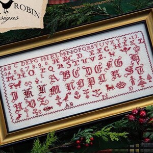 May include: Framed cross-stitch sampler with a gold-colored frame. The sampler features a red alphabet, numbers, and decorative motifs on a white background. The text "LITTLE ROBIN designs" is in the upper left corner.