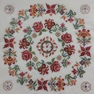 May include: A white fabric with a cross-stitch design featuring a clock surrounded by flowers and leaves. The clock face is white with black Roman numerals and hands. The flowers are red, orange, and green. The leaves are green. The fabric is white with a subtle texture. The design is framed by a border of small, red, orange, and green flowers.