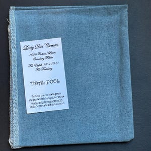 May include: A square, light blue fabric piece with a white tag. The tag reads "Lady Dot Creates" and lists fabric details: 100% Cotton/Linen, Oinabang Fabric, Fat Eighth 18" x 10.5". The text "TIDAL POOL" is also visible.