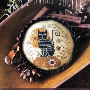May include: A black cat cross-stitch design in a round, brown, metal frame. The cat is sitting on a pumpkin with a moon and stars in the background. The design is titled "Olga's Tart" and is from "Jack's Sweet Shoppe".