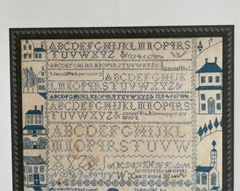 May include: A framed sampler with the text "Ann McFarlan Sampler 1827." The sampler features embroidered letters, numbers, and small illustrations of houses and trees in blue thread on a cream-colored background. The frame is dark.