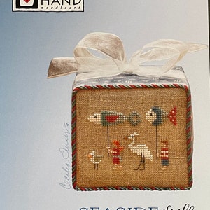 Puede incluir: Un patr&oacute;n de punto de cruz para un adorno con tem&aacute;tica mar&iacute;tima. El patr&oacute;n presenta un p&aacute;jaro blanco, dos ni&ntilde;os y dos peces sosteniendo banderas. El patr&oacute;n se titula "Seashore Frill" y es de Heart in Hand needleart.