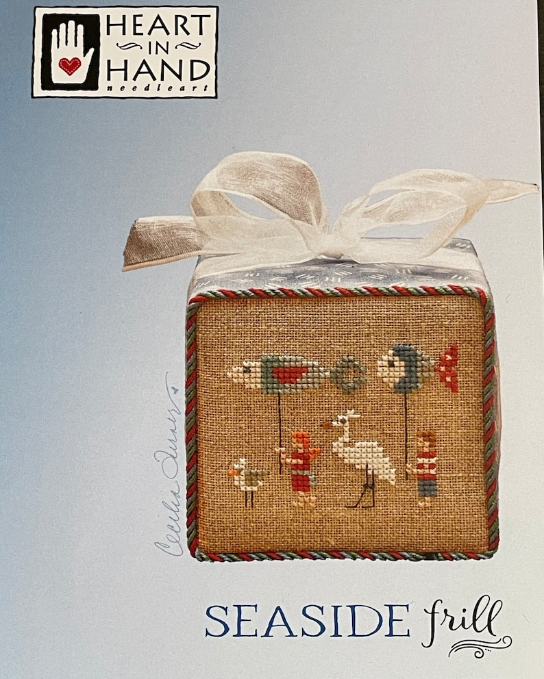 Puede incluir: Un patr&oacute;n de punto de cruz para un adorno con tem&aacute;tica mar&iacute;tima. El patr&oacute;n presenta un p&aacute;jaro blanco, dos ni&ntilde;os y dos peces sosteniendo banderas. El patr&oacute;n se titula "Seashore Frill" y es de Heart in Hand needleart.