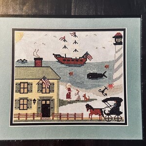 May include: A cross-stitch embroidery depicting a seaside scene with a yellow house, a red sailboat, a whale, a lighthouse, and a horse-drawn carriage. The scene is framed in a light blue mat and a black frame.