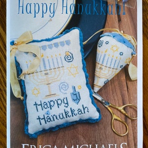 May include: A cross-stitch pattern for a pillow with the text "Happy Hanukkah" and a menorah design. The pattern is from the "The Buttoned Up Collection" by Erica Michaels Needleart Designs.