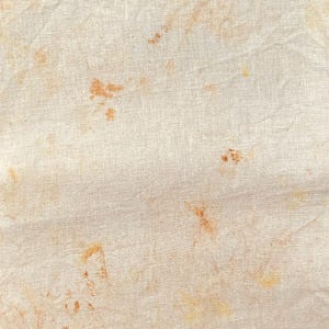 May include: A light beige fabric with a visible woven texture featuring scattered, irregular splotches of warm orange and golden yellow pigment. The surface displays a natural, organic appearance with subtle creases and a consistent, fibrous weave.