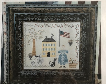 Pode incluir: Um padrão de ponto de cruz intitulado "Liberty" de Samplers Not Forgotten. O design apresenta uma casa, uma árvore, uma bicicleta, um cão, um pássaro e uma bandeira americana.