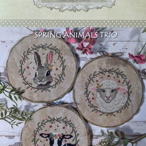 May include: Three cross stitch patterns featuring a bunny, a lamb, and a cow, each wearing a floral crown and surrounded by a wreath of leaves and flowers. The patterns are titled "Spring Animals Trio" and are by "puntiimpuntini".