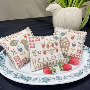 May include: Three embroidered cushion covers with a summer theme. The cushions are white with a light brown background and feature a variety of designs, including a bird, a house, and a strawberry plant. The cushions are decorated with a lace trim and the text "Hello Summer" is embroidered on one of the cushions.