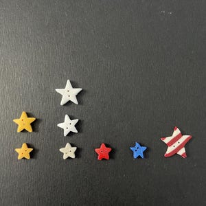 May include: A collection of star-shaped buttons in various colors, including yellow, white, red, and blue. One button features a red and white striped design. The buttons are arranged on a dark surface.
