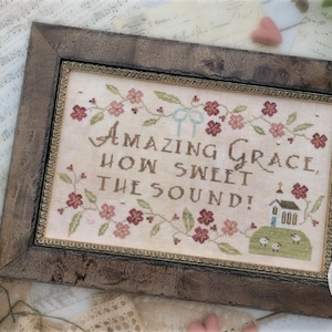 May include: Framed cross-stitch sampler with the text "Amazing Grace, How Sweet the Sound!" in brown thread. The sampler features floral and church motifs, framed in a rustic wooden frame. The design is by Brenda Gervais.