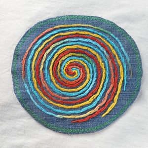 May include: Circular denim patch with a spiral design embroidered in vibrant colors: red, orange, yellow, turquoise, and blue. The patch has a green stitched border and is set against a white background.