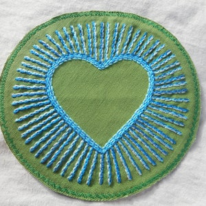 May include: A round, green embroidered patch with a blue heart in the center. The heart is surrounded by radiating lines of blue and white thread. The patch has a green stitched border.