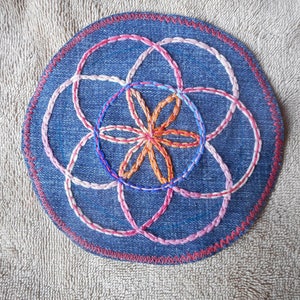 May include: Round denim patch with an embroidered geometric design. The design features overlapping circles and a central flower shape, stitched in white, pink, orange, and blue thread. The patch has a red stitched border.