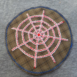 May include: A round, brown patch with a plaid pattern, featuring a white and red embroidered design resembling a spider web. The patch has a blue stitched border and is set against a gray background.