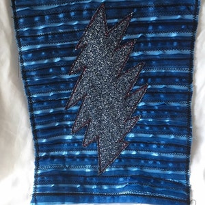 May include: A blue fabric patch with a lightning bolt design. The background is a textured blue with horizontal stripes. The lightning bolt is a darker blue with a floral pattern and red stitching.