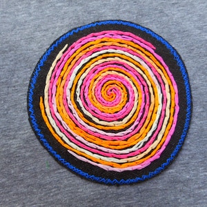 May include: A gray t-shirt featuring a circular embroidered patch. The patch has a black background with a blue stitched border. Inside, a spiral design uses pink, orange, and white threads.
