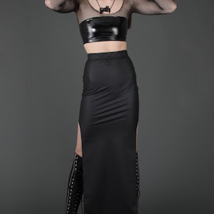 May include: A black long-sleeved shirt with a mesh panel and a black patent leather crop top. The model is wearing a black skirt with a side slit and black knee-high boots. The model is wearing a necklace with the word "Goth" on it.