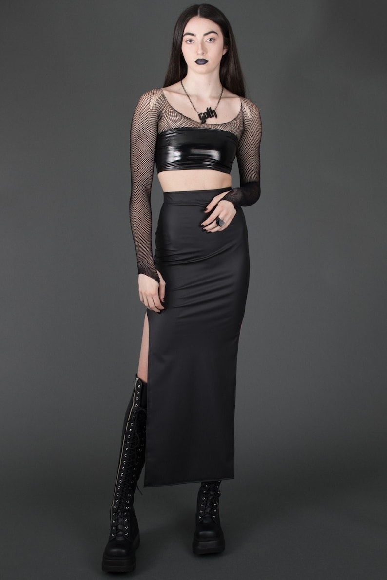 May include: A black long skirt with a side slit and a black fitted top with a black fishnet long-sleeve shirt underneath. The model is wearing a black necklace with the word "Goth" on it.