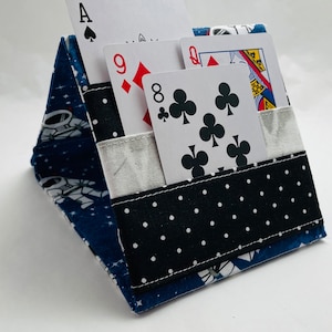 May include: A blue and white fabric card holder with a space theme. The holder has a pocket for cards and a flap that folds over to keep the cards secure. The holder is decorated with white stars and astronauts on a blue background. The holder is shown with four playing cards: the Ace of Spades, the 9 of Diamonds, the 8 of Clubs, and the Queen of Hearts.