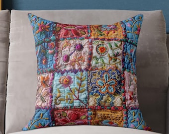 May include: A square decorative pillow with a patchwork design. The pillow features a variety of colorful embroidered squares with floral and geometric patterns. The color palette includes blues, reds, oranges, and greens. The pillow is placed on a light gray sofa.