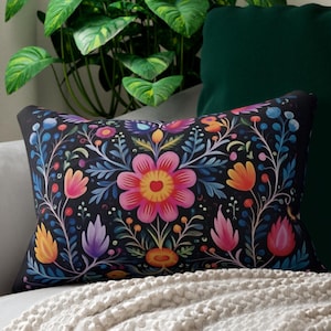 May include: A decorative rectangular pillow with a black background and a vibrant floral pattern. The design features a large pink flower at the center, surrounded by colorful blooms in shades of orange, blue, and purple. The pillow is placed on a gray sofa.