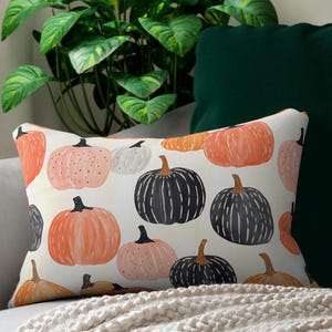 May include: Rectangular cushion with a pumpkin pattern in orange, pink, white, and black on a cream background. The cushion is on a grey sofa with a cream throw. The scene includes a green plant and a dark green cushion.
