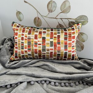 Puede incluir: Una almohada rectangular con un patr&oacute;n geom&eacute;trico en tonos rojos, amarillos y marrones. La almohada est&aacute; sobre una manta gris con pompones alrededor del borde.