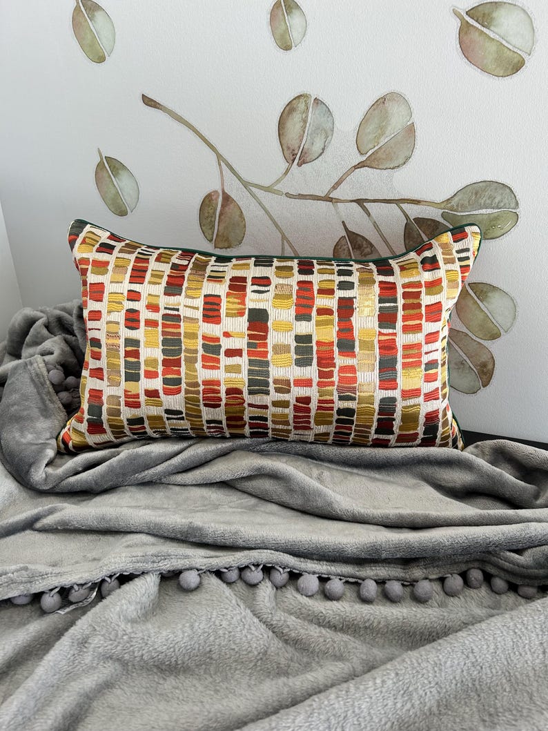 Puede incluir: Una almohada rectangular con un patr&oacute;n geom&eacute;trico en tonos rojos, amarillos y marrones. La almohada est&aacute; sobre una manta gris con pompones alrededor del borde.